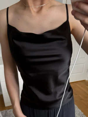 Svart satinlinnetopp från Gina Tricot - Säljer en svart linnetopp i glansigt satinmaterial från Gina Tricot. Toppen har tunna axelband och något lös passform. Perfekt att styla med jeans eller kjol för en fest eller vardags look.