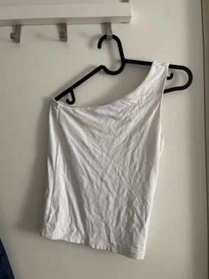 Vit oneshoulder topp - Snygg vit oneshoulder topp i mjukt material. Perfekt för dig som vill ha en enkel men trendig look. Toppen är ärmlös och har en asymmetrisk skärning över axeln.