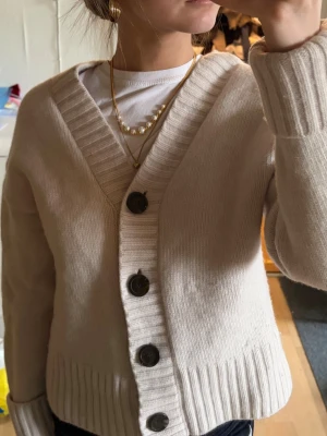 Beige ribbad kofta med stora knappar - Mysig beige kofta med djup V-ringning och stora mörka knappar framtill. Perfekt att slänga över en t-shirt för en enkel och snygg look. Endast använd fåtal gånger och är i bra skick. Materialet är Cashmere och Wool💕💕😇