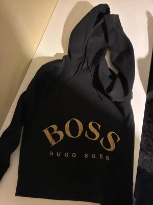Svart hoodie från Hugo Boss - Snygg svart hoodie från Hugo Boss med stor guldfärgad BOSS-logga på bröstet. Klassisk modell med huva och dragsko. Tillverkad i mjukt material som känns skönt mot huden. Perfekt för dig som gillar stilrena och trendiga plagg.