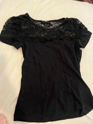 Svart topp med spetsdetalj från H&M - Snygg svart topp från H&M med spets över axlar och bröst. Kortärmad modell i mjuk bomull med rund halsringning och transparent spets som ger en extra touch. Perfekt att styla med jeans eller kjol för en trendig look. perfekt för sommaren men också fin på vintern