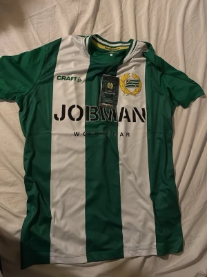 Hammarby matchtröja från Craft - Grön och vit randig Hammarby matchtröja från Craft med klubbmärke på bröstet och JOBMAN Workwear som sponsortryck. Tillverkad i lätt och ventilerande funktionsmaterial, perfekt för fotboll eller träning. Klassisk rund hals och korta ärmar.