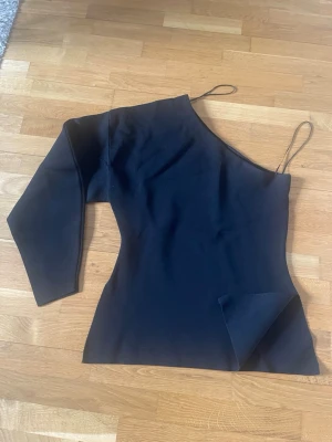 Svart oneshoulder topp - Trendig svart topp med oneshoulder-design från Zara stl.s. Toppen har lång ärm på ena armen och ärmlös på andra, vilket ger en cool asymmetrisk look. Perfekt för dig som vill sticka ut med en unik stil.