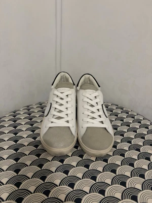 Vita sneakers från Philippe Model - Stilrena vita sneakers från Philippe Model med detaljer i svart och grå mocka på tån. Skorna har vita snören, svart hälparti och en ikonisk sköldlogga på sidan. Tillverkade i läder och mocka för en lyxig känsla och modern look.