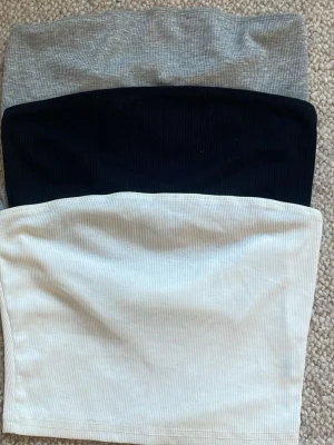 3-pack tubtoppar - Säljer ett set med tre ribbade tubtoppar i färgerna grå, svart och vit. Perfekta att styla till jeans eller kjol för en enkel och clean look. Alla topparna är axelbandslösa och har en mjuk, stretchig passform.