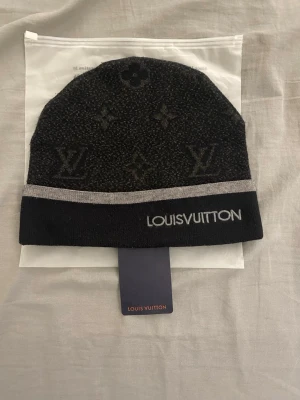 Svart mössa  - Exklusiv svart mössa från med klassiskt monogrammönster och logotyp broderad på uppviket. Mössan har en diskret grå rand och är stickad i mjukt material som ger en lyxig känsla. Perfekt för dig som vill ha en stilren accessoar med premiumkänsla.