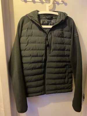 Mörkgrå vindjacka med huva Helly Hansen - Snygg mörkgrå vindjacka från Helly Hansen med quiltad front och rygg, samt mjuka ärmar. Jackan har huva, dragkedja framtill och en bröstficka med dragkedja. Perfekt för blåsiga dagar och enkel att matcha till olika outfits. Priset går att diskutera och jackan kommer med original taggen.