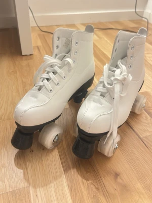 Vita klassiska rullskridskor - Säljer ett par vita rullskridskor med högt skaft och snörning. De har fyra hjul per sko och svart tåstopp. Skorna är tillverkade i syntetmaterial och har en retro look som passar perfekt för dans eller cruising på gatorna. säljer de för att jag inte använder de  (använts bara 3 gånger). 