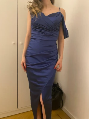 Blå axelbandsklänning med slits - Snygg blå långklänning med smala axelband och draperad överdel. Klänningen har en slits framtill och figurnära passform som framhäver siluetten. Tillverkad i ett glansigt, satängliknande material som ger en lyxig känsla.