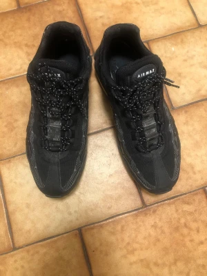 Nike Air Max 95 svarta sneakers - Nike Air Max 95 sneakers i helsvart med vita detaljer och reflekterande mönster längs sidorna. Skorna har en chunky sula med synlig Air-enhet, mesh och syntetmaterial i ovandelen samt svarta snören med vita inslag. Perfekt för dig som gillar streetwear och sportig stil.