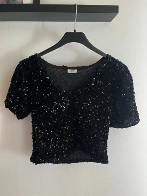 Svart glittrig croppad topp JDY - Säljer en svart croppad topp från JDY med korta ärmar och massor av glittrande paljetter. Toppen har en v-ringad hals och är perfekt för dig som vill sticka ut. Materialet känns mjukt och följsamt mot huden.