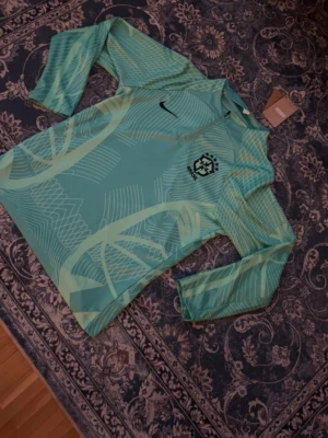 Nike Brasil långärmad träningströja - Snygg långärmad träningströja från Nike i ljus turkos med grafiskt mönster och Brasilien CBF-logga på bröstet. Tröjan har dragkedja vid halsen och är tillverkad i ett lätt, tekniskt material som passar perfekt till fotbollsträning.