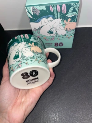 Muminmugg 80 år Arabia jubileumsmugg - Muminmugg från Arabia med 80-årsjubileumsmotiv. Muggen är i keramik och har ett färgglatt tryck med Muminfigurer i grönt, vitt, rosa och orange. Levereras i originalförpackning och streckkod på, med matchande design. Perfekt för samlare