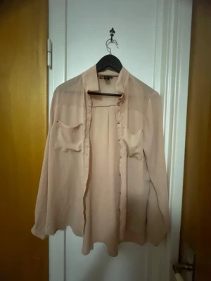 Beige transparent blus med fickor - Säljer en beige, transparent blus med långa ärmar och två bröstfickor. Blusen har knappar framtill och en lös, flowy passform. Perfekt att styla lager på lager. Materialet känns lätt och luftigt.