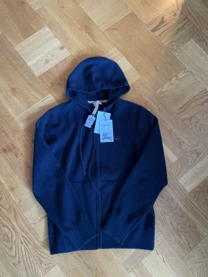 Mörkblå hoodie från Burberry - Snygg mörkblå hoodie från Burberry med dragkedja framtill och klassisk huva. Hoodien har Burberry-logga broderad på bröstet och är tillverkad i mjukt material som känns lyxigt. Perfekt för dig som vill ha en stilren och exklusiv look.