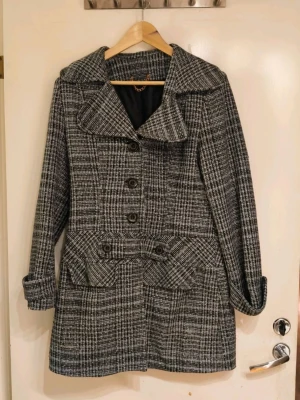 Grå-rutig rock med stora knappar - Ch@nel-inspirerad snygg grå-rutig rock med stora svarta knappar och bred krage. Rocken har två stora fickor framtill med lock och dekorativa knappar, samt ett bälte i midjan baktill. Materialet ser ut att vara ullblandning och fodrad insida. Perfekt för kyligare dagar.