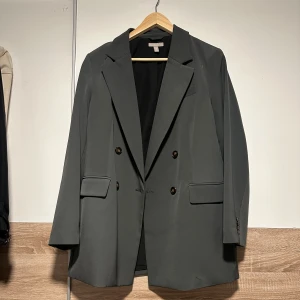 Mörkgrå oversized kavaj - Stilren mörkgrå kavaj med dubbelknäppning och breda slag. Kavajen har två framfickor med lock och en klassisk rak passform. Perfekt för en trendig och avslappnad look. Fodrad insida för extra komfort.