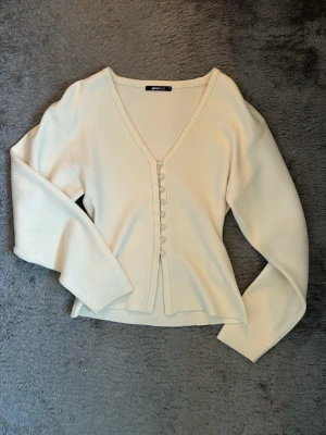 Krämvit kofta från Gina Tricot - Säljer en krämvit kofta från Gina Tricot med v-ringning och dekorativa knappar framtill. Koftan har långa ärmar och är tillverkad i ett mjukt, tunt stickat material. Har aldrig använts och ser helt ny ut💖