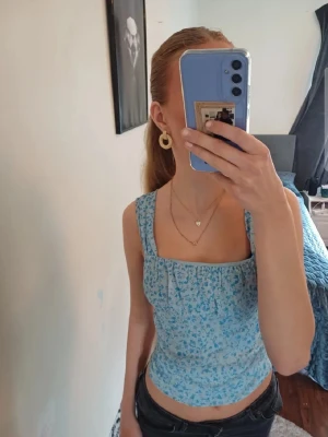 Blommig blå linnetopp - Söt blå linnetopp med blommigt mönster och breda axelband. Toppen har en smickrande, något tight passform och fyrkantig ringning både fram och bak. 