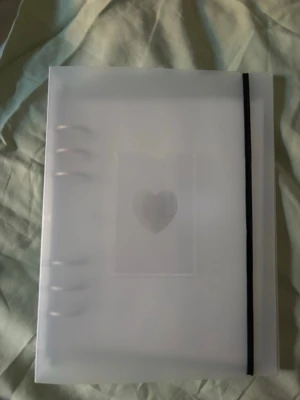 Genomskinligt samlaralbum med hjärta - Säljer ett genomskinligt samlaralbum i plast med svart resårband och hjärtmotiv på framsidan. Inuti finns plastfickor för att förvara kort, bilder eller små samlarobjek, brukar oftast användas för photocards. Perfekt för dig som vill organisera och visa upp din samling på ett snyggt sätt.