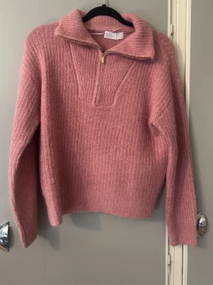 Rosa stickad tröja med halv dragkedja - Mysig rosa stickad tröja från Samsøe Samsøe med hög krage och halv dragkedja framtill. Tröjan har en avslappnad passform, långa ärmar och är gjord i ett fluffigt material som känns riktigt skönt. Perfekt för kalla dagar och enkel att styla.