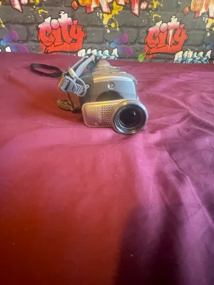 Silverfärgad digital videokamera - Säljer en kompakt digital videokamera i silver med utfällbar skärm och handledsrem. Kameran har en klassisk design med tydliga knappar och lins framtill. Perfekt för dig som vill filma och spara minnen digitalt.