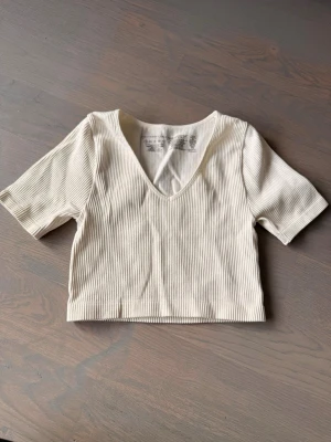 Ribbad vit croppad topp - Säljer en ribbad vit croppad topp med v-ringning och korta ärmar. Toppen har en enkel och stilren design som passar perfekt till jeans eller kjol. Materialet är mjukt och stretchigt för en bekväm passform.