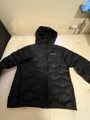 Svart dunjacka Peak Performance - Snygg svart dunjacka från Peak Performance med huva och dragkedja framtill. Jackan har ett quiltat mönster och är tillverkad i ett glansigt material som ger en modern look. Perfekt för kalla vinterdagar och riktigt varm. MAN