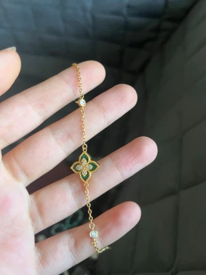 Guldigt armband med grön detalj - Snyggt armband i guldfärgad metall med en fyrklöverformad detalj i mitten, dekorerad med gröna och vita stenar. Länken har små pärlliknande stenar längs kedjan som ger extra bling. Perfekt accessoar för att lyfta din outfit.