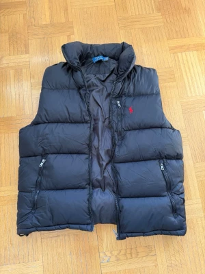 Svart dunväst från Polo Ralph Lauren - Snygg svart dunväst från Polo Ralph Lauren med hög krage och dragkedja framtill. Västen har två sidofickor med dragkedja och en bröstficka, samt den klassiska röda loggan broderad på bröstet. Perfekt för lager-på-lager och riktigt skön att bära. Använd fåtal gånger. Storleken är i L. Hör av er vid frågor☺️