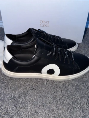 Svarta sneakers från Oliver Cabell - Snygga svarta sneakers från Oliver Cabell med vit sula och vit detalj på sidan. Skorna har svarta skosnören med logga och vit hälkappa. Tillverkade i slätt läder för en clean look. Perfekta för dig som gillar stilrena och moderna sneakers. Skick 10/10. Använda en gång. Allt og ingår. Hör av dig vid frågor!😊 (använda 1 gång)