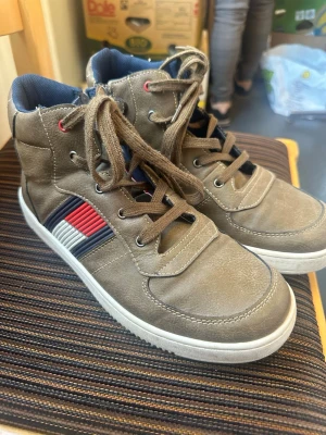  Tommy Hilfiger bruna high top sneakers med ränder - Snygga bruna high top sneakers med snörning och vita sulor. Sidan har coola detaljer i form av randiga inslag i rött, vitt och blått. Insidan är fodrad med mörkblå textil och ovandelen är i syntetmaterial. Perfekta för dig som vill ha en stilren men ändå unik look.