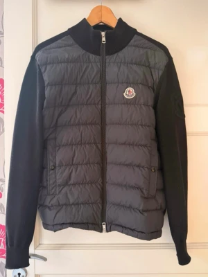 Svart pufferjacka från Moncler - Snygg svart pufferjacka från Moncler med quiltad front och stickade ärmar. Jackan har dragkedja framtill, två fickor med knapp och Moncler-logga på bröstet samt ett broderat emblem på ärmen. Perfekt för kalla vinterdagar och riktigt trendig look.