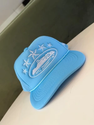 Blå truckerkeps med broderade stjärnor - Säljer en ljusblå truckerkeps med vit brodyr framtill och på sidan. Kepsen har ett broderat motiv av en bil och flera stjärnor. Baksidan är i mesh för extra ventilation och kepsen har böjd skärm. Perfekt för dig som vill sticka ut med en unik accessoar.