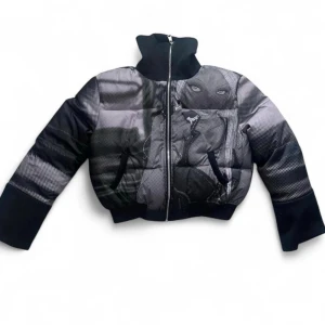 Svart pufferjacka med grafiskt tryck - Unik svart pufferjacka med hög krage och dragkedja framtill. Jackan har ett stort grafiskt tryck i gråskala med ett ansikte och konstnärliga detaljer på både fram- och baksida. Oversized passform och breda ärmar ger en cool streetwear-look. Perfekt för dig som vill sticka ut.