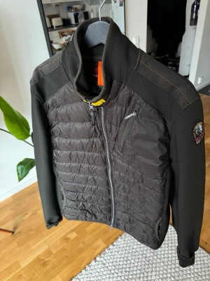 Olivgrön hybridjacka från Parajumpers - Snygg olivgrön hybridjacka från Parajumpers med quiltad front och meshdetaljer på ärmar och axlar. Jackan har dragkedja framtill, bröstficka med zip och patch på ärmen. Perfekt för dig som vill ha en sportig och modern look.
