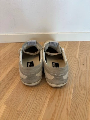 Golden Goose vita låga sneakers - Snygga vita sneakers från Golden Goose med ikonisk stjärndetalj på sidan och blå logga på plösen. Skorna har en vintage look, vita skosnören och är tillverkade i mocka och läder. Perfekta för dig som gillar streetstyle och vill ha något unikt.