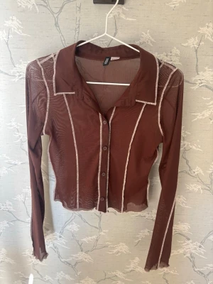 Brun meshblus med kontrastsömmar - Trendig brun meshblus med långa ärmar och vita kontrastsömmar. Blusen har krage, knäppning framtill och figurnära passform. Perfekt för lager-på-lager och en cool, modern look.