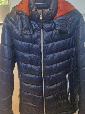 Mörkblå dunjacka från Jack & Jones - Snygg mörkblå dunjacka från Jack & Jones med orange innerfoder och justerbar huva. Jackan har dragkedja framtill, två fickor med dragkedja och vattenavvisande material. Perfekt för kalla vinterdagar och riktigt skön passform.