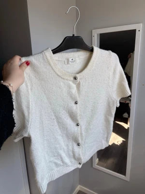 Vit kortärmad kofta från H&M - Söt vit kofta från H&M med korta ärmar och rund halsringning. Koftan har ribbade muddar och knäpps med silverfärgade knappar framtill. Perfekt att slänga över en topp för en enkel och clean look. Är lite nopprig men i övrigt super fint skick❣️
