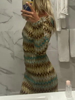 Mönstrad långärmad klänning - Säljer en figurnära klänning med långa ärmar och zigzag-mönster i beige, brunt, turkost och guld. Säljer då den inte kommit till användning, därav är den i bra skick! Kontakta vid fler frågor.