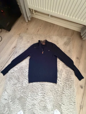 Mörkblå halvzip-tröja från Morris  - Snygg mörkblå stickad tröja från Morris  med halv dragkedja och liten logga broderad på bröstet. Tröjan har ribbade muddar och krage med kontrastfoder. Perfekt för lager på lager och stilren look. Storlek xs 