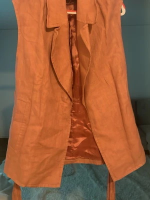 Rosa lång väst med skärp - Stilren väst i längre modell med bred krage och knytband i midjan. Västen har slät yta och är fodrad med glansigt tyg på insidan. Perfekt för lager på lager och ger en trendig look till din outfit.