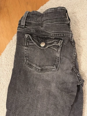 Gråa bootcut jeans med fickdetalj - Säljer ett par gråa jeans med bootcut passform och snygg ficka med knapp bak. Jeansen har klassisk femficksdesign och är i ett mjukt bomullsmaterial. Perfekta till en avslappnad stil och passar till det mesta. Storlek 158 men passar xs! Passade mig när jag var ca 161!! De är använda men det enda som är lite slitet är att fickorna har böjts upp lite (se bild 1)