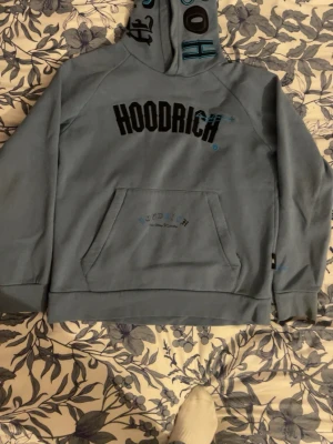 Blå hoodrich hoodie L - Säljer en blå hoodie från Hoodrich med svart och blått tryck på bröstet och detaljer på huvan. Hoodien har en klassisk känguruficka framtill och ribbade muddar vid ärmslut och nederkant. Perfekt för dig som gillar streetwear och vill ha något bekvämt och stilrent.