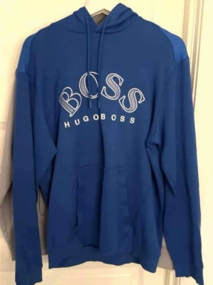 Blå hoodie från Hugo Boss - Snygg blå hoodie från Hugo Boss med stor vit BOSS-logga och text på bröstet. Hoodien har huva med dragsko och en klassisk magficka framtill. Tillverkad i mjukt material som känns skönt mot huden. Perfekt för en avslappnad och trendig look.