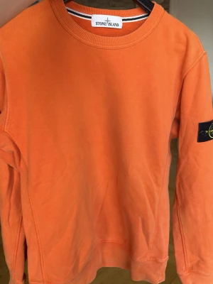 Orange sweatshirt från Stone Island - Säljer en orange sweatshirt från Stone Island med rund hals och den klassiska svarta patchen på vänster ärm. Tröjan är gjord i mjuk bomull och har ribbade muddar vid ärmslut och nederkant. Perfekt för dig som gillar streetwear och vill ha något som sticker ut. Säljer eftersom den är för liten