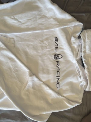 Vit hoodie från Sail Racing - Säljer en vit hoodie från Sail Racing med svart logga och text på bröstet. Hoodien har huva med dragsko och en klassisk känguruficka framtill. Tillverkad i mjukt bomullsmaterial som är skönt att ha på sig. Perfekt för dig som gillar stilren och sportig design.