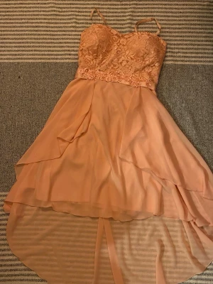 Ljusrosa spetsklänning med volang - Ljusrosa klänning med spetsliv, tunna axelband och omlott volangkjol. Livet har detaljer i spets och broderade blommor på satinkant. Kjolen är asymmetrisk och gjord i ett lätt, flygande material som ger en elegant look.