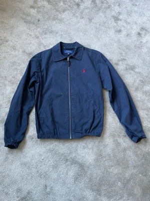Marinblå vindjacka från Polo Ralph Lauren - Klassisk marinblå vindjacka från Polo Ralph Lauren med dragkedja framtill, två sidofickor och elastisk midja samt ärmslut. Jackan har en röd broderad logga på bröstet och är tillverkad i ett lätt bomullsmaterial. Perfekt för en stilren och avslappnad look.